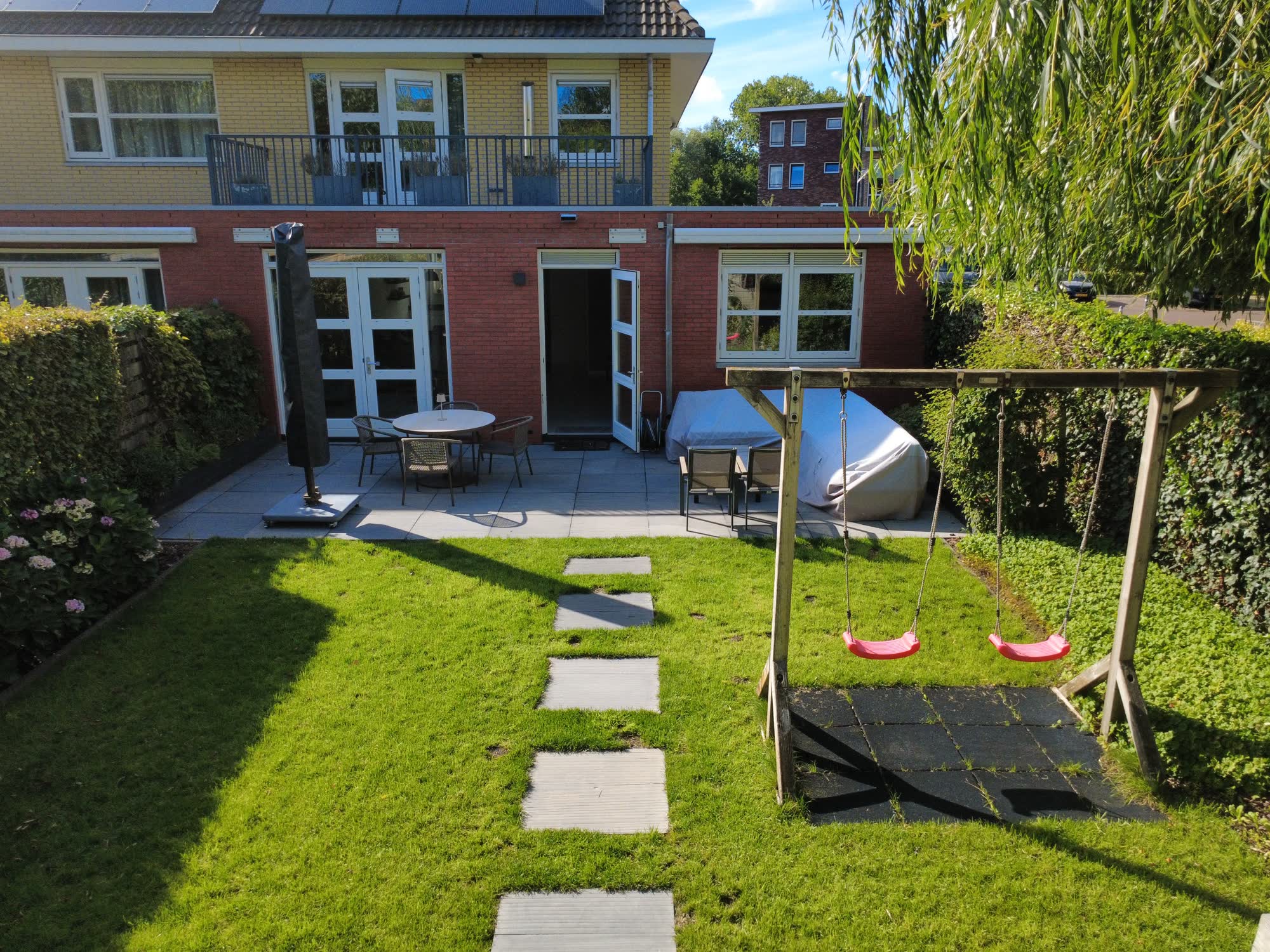 De achtertuin met terras, tuinset en schommel, met het huis erachter