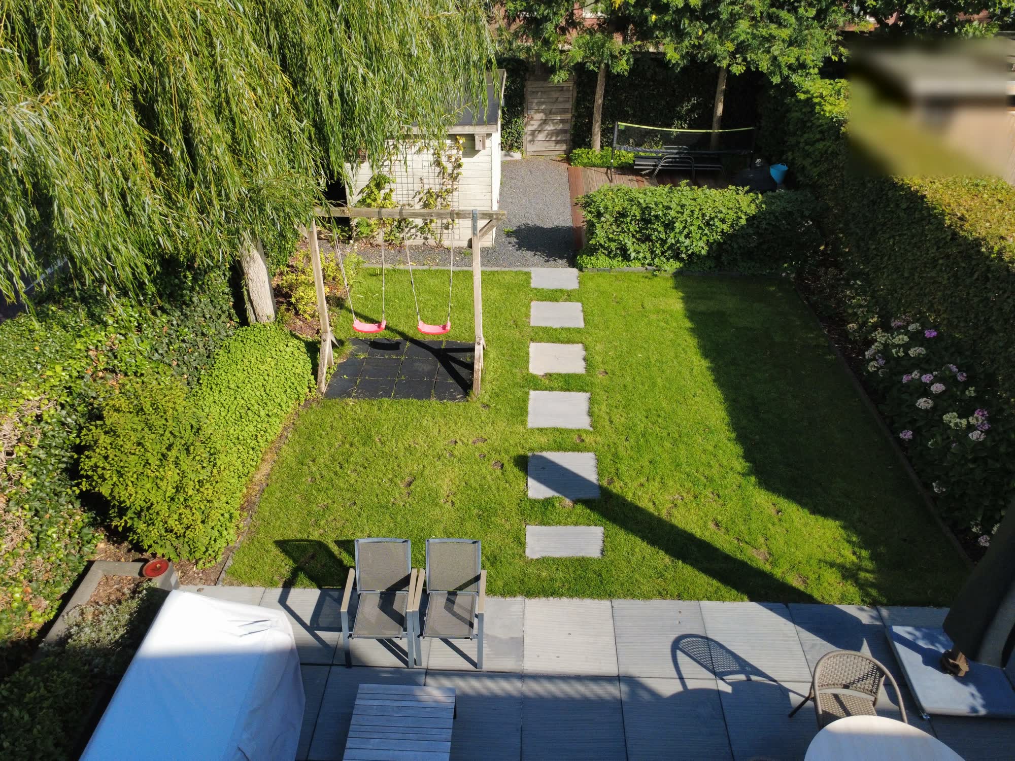 De hele achtertuin van bovenaf met gras, terras, schommel en tuinhuis